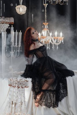 Uzun kızıl saçlı, siyah elbiseli, zarif bir odada kristal avizelerle poz veren güzel bir kadın.