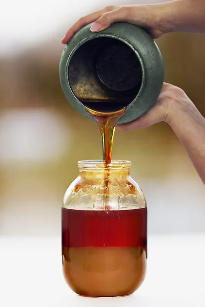 Woman's hands pours a honey from a pot to a jar — Stock Photo, Image