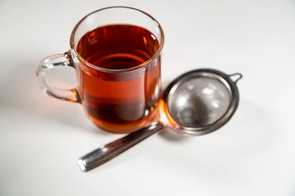 Beyaz bir masanın üzerinde gümüş paslanmaz çelik çay süzgeci olan kupada temiz cam bir bardak Seylan siyah çayı.