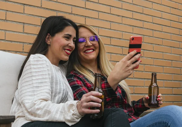İki gülümseyen genç arkadaş bira içiyor ve terasta cep telefonuyla selfie çekiyor.