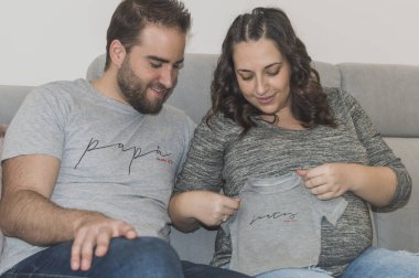 Hamile düğün çifti kanepede oturup birbirlerine bakıp gülümsüyor.