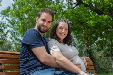 Hamile çift bankta oturuyor ve parkta poz veriyor.