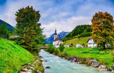 Geniş panoramik manzara ikonik St. Sebastian Kilisesi 'ni, Ramsauer Ache Nehri' ni ve renkli sonbahar ağaçlarıyla çerçevelenmiş dağlık köy evlerini yakalar. Ramsau bei Berchtesgaden, Almanya