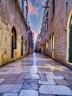 Düşük açılı bir çekim, gün batımından sonra dar bir caddenin parlayan kaldırım taşlarına yansıyan, dükkan ışıklarının sıcak parıltısını yakalar. Dubrovnik, Hırvatistan