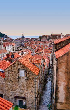 A vertical shot from the city walls looking down into a narrow cobblestone street lined with red-roofed houses under a clear summer sky. Dubrovnik, Croatia
