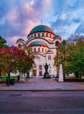 Saint Sava Kilisesi 'nin ana girişinin dikey günbatımı görüntüsü ve sonbahar ağaçları tarafından çerçevelenmiş güneş ışığıyla aydınlatılmış kubbesi. Belgrad, Sırbistan