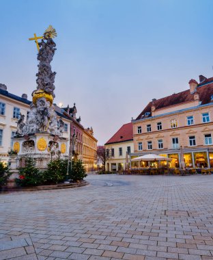 Avusturya 'da alacakaranlıkta Baden bei Wien' in sakin ana meydanındaki Kutsal Üçlü Sütun 'un dikey görüntüsü ve aydınlatılmış tarihi binalar.