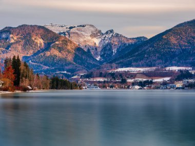 Avusturya 'da kışın arka planda güneşli karla kaplı dağlarla Wolfgang Gölü' nün karşısındaki St. Wolfgang köyünün manzarası uzun süre görülür.