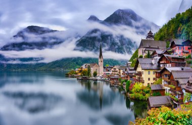 Avusturya 'daki dağ sisinin altındaki gölün ipeksi yumuşak sularını yansıtan Hallstatt' ın dik dağlarına tünemiş geleneksel evlerin panoramik manzarası.