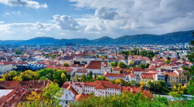 Avusturya 'da bulutlu bir gökyüzü altında sonbahar ağaçları arasında tarihi kırmızı çatılar ve modern mavi Kunsthaus sanat müzesinin yer aldığı Graz manzaralı bir şehir.