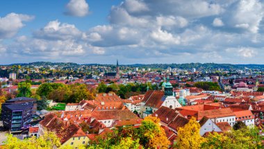 Avusturya 'da bulutlu mavi bir gökyüzünün altında Herz-Jesu-Kirche kulesinin ve sonbahar ağaçlarının arasında tarihi kırmızı çatıların yer aldığı Graz Eski Kasabasının manzaralı yüksek açılı manzarası.