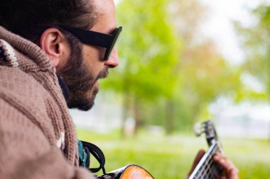 Sakallı ve gözlüklü hippi çimlerin üzerinde gitar çalıyor.