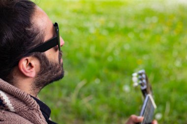 Sakallı ve gözlüklü hippi çimlerin üzerinde gitar çalıyor.