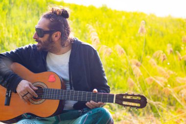 Sakallı ve gözlüklü hippi çimlerin üzerinde gitar çalıyor.