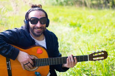 Sakallı ve gözlüklü hippi çimlerin üzerinde gitar çalıyor.