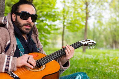 Sakallı ve gözlüklü hippi çimlerin üzerinde gitar çalıyor.