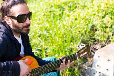 Sakallı ve gözlüklü hippi çimlerin üzerinde gitar çalıyor.