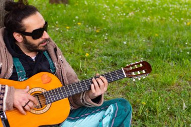 Sakallı ve gözlüklü hippi çimlerin üzerinde gitar çalıyor.