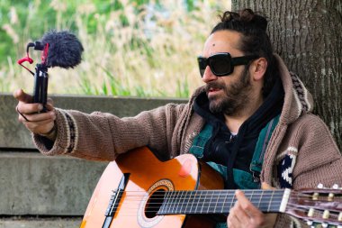 Sakallı, gözlüklü, gitar çalan hippi adam dışarıda çimlerin üzerinde video çekiyor.