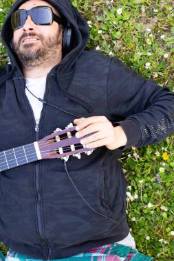 Hippi sakallı ve gözlüklü bir adam çimlerin üzerinde gitar çalıyor.