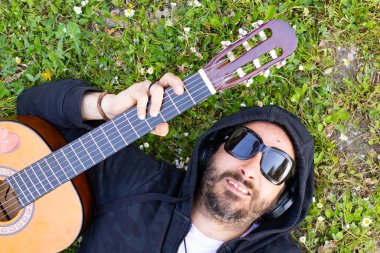 Hippi sakallı ve gözlüklü bir adam çimlerin üzerinde gitar çalıyor.