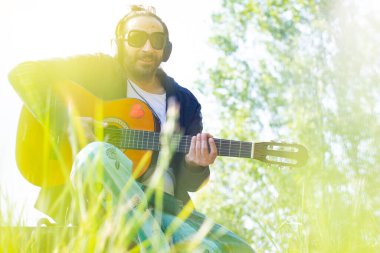 Sakallı ve gözlüklü hippi çimlerin üzerinde gitar çalıyor.