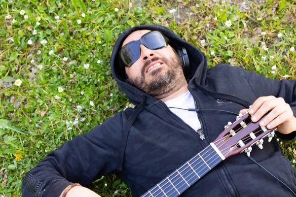 Hippy man lying down with beard and glasses playing guitar outdoors on the grass