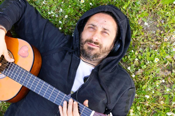 Hippi sakallı ve kulaklıklı bir adam çimlerin üzerinde gitar çalıyor.