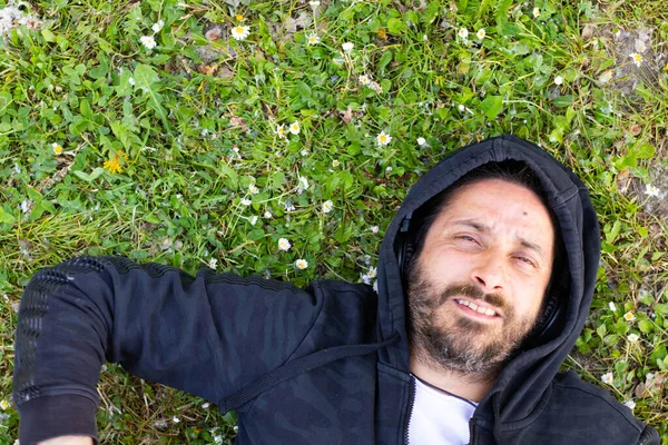 Hippy man lying down with beard and headphones playing guitar outdoors on the grass