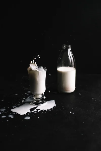 Une bouteille de lait rustique et un verre de lait sur fond noir avec éclaboussure liquide de lait — Photo