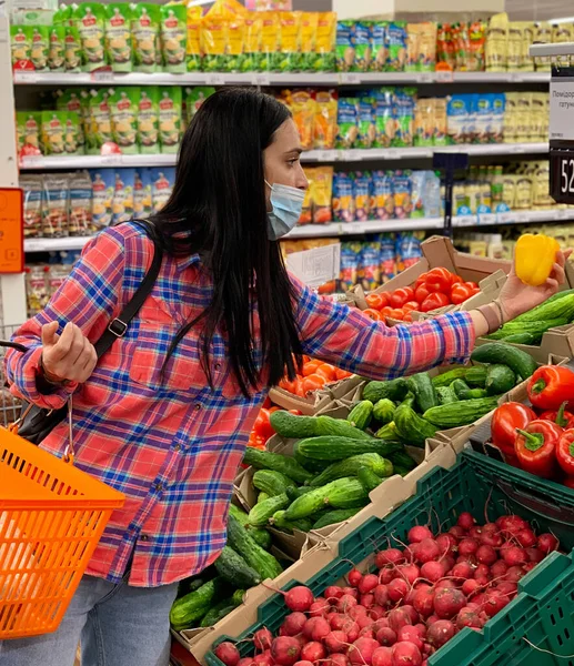 Woman in a medical mask choosing vegetables in a grocery supermarket. Protection from the coronavirus epidemic, increased immunity with fresh fruit. Healthy food for fighting viruses.
