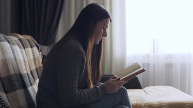 A young woman sitting on a comfortable sofa in a cozy living room, reading a book and relaxing at home. Concept of peaceful leisure, weekend rest, and self-care moments. Warm soft light creates a calm and inviting atmosphere.