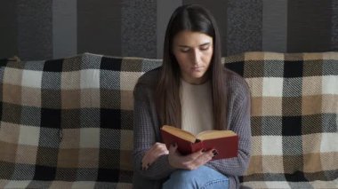 A young woman sitting on a comfortable sofa in a cozy living room, reading a book and relaxing at home. Concept of peaceful leisure, weekend rest, and self-care moments. Warm soft light creates a calm and inviting atmosphere.