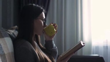 A young woman sitting comfortably on a sofa at home, holding a cup of hot coffee or tea while reading a book. Cozy home atmosphere, relaxation, weekend leisure, mindfulness, and peaceful lifestyle concept. Soft natural light