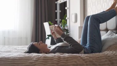 Genç bir kadın yatakta uzanıyor ve parlak bir yatak odasında kitap okuyor. Huzurlu bir iç atmosfer, rahatlama ve kişisel bakım anı.