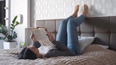 Genç bir kadın yatakta uzanıyor ve parlak bir yatak odasında kitap okuyor. Huzurlu bir iç atmosfer, rahatlama ve kişisel bakım anı.