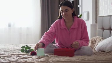 Sevgililer Günü sabahı yatağında romantik bir hediyeyle oturan genç bir kadın. Hediyeye iliştirilmiş bir notu okuyor ve bir buket çiçeğin tatlı bir gülümsemeyle kokusunu alıyor. Sıcak ve samimi ev atmosferi, aşk, romantizm ve şefkat kavramı