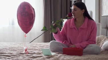 Sevgililer Günü sabahı yatağında romantik bir hediyeyle oturan genç bir kadın. Hediyeye iliştirilmiş bir notu okuyor ve bir buket çiçeğin tatlı bir gülümsemeyle kokusunu alıyor. Sıcak ve samimi ev atmosferi, aşk, romantizm ve şefkat kavramı