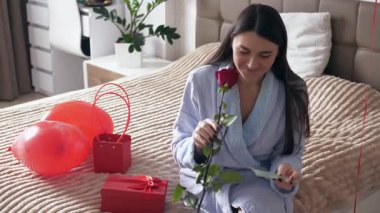 Sevgililer Günü sabahı yatağında romantik bir hediyeyle oturan genç bir kadın. Hediyeye iliştirilmiş bir notu okuyor ve bir buket çiçeğin tatlı bir gülümsemeyle kokusunu alıyor. Sıcak ve samimi ev atmosferi, aşk, romantizm ve şefkat kavramı