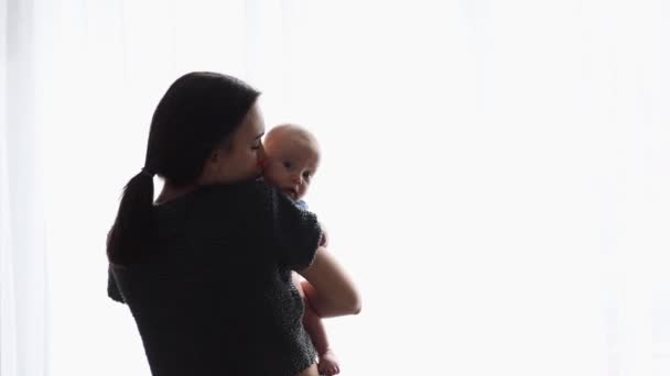 Anne pencerenin ışığında bebeğini kucaklıyor ve öpüyor. — Stok video