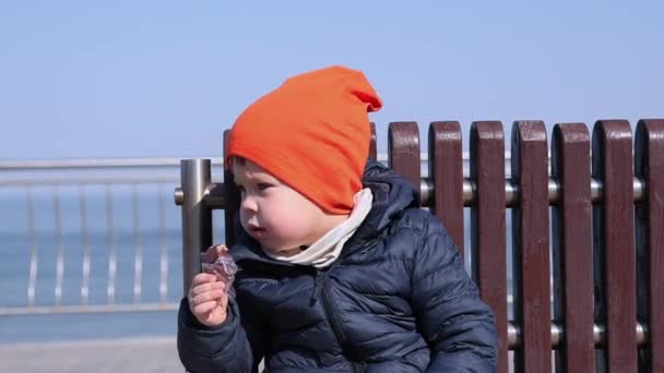 Malý chlapec sedí na lavičce v moři a jí čokoládu — Stock video