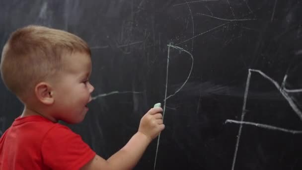 Criança menino miúdo em vermelho camisa escreve no quadro negro — Vídeo de Stock