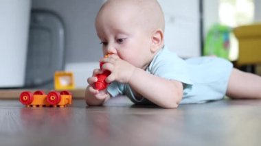 6 aylık küçük bebek, tahta zeminde karnının üstünde yatıyor oyuncaklarla oynuyor.