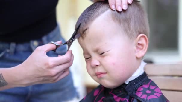 Retrato de un niño pequeño con los ojos cerrados y emociones aterradoras mientras la peluquera corta su flecos con tijeras — Vídeo de stock