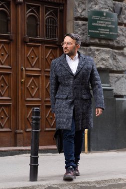 Adam sokağa dökülüyor. Gri saçlı, gözlüklü, beyaz gömlek, ceket ve koyu renk ceket giyen bir adam. Güneşli gün dışarıda. Orta yaşlı bir adam sokakta ince zekayla yürüyor.