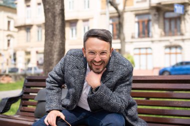 Parkta oturan adam kahverengi bir bankta oturuyor ve gülümsüyor. Baharda, yeşil çimenler, güneş parlıyor, hava güzel. şehir arka planında. Gözlüklü, beyaz gömlekli ve gri ceketli genç adam.