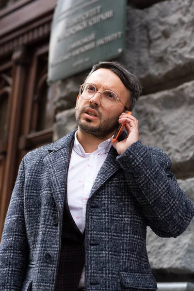 Man are talking on the phone on the street. A man with gray hair and a beard wearing glasses, wearing a white shirt, jacket and dark coat. holds a phone in his hands