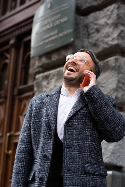 Man are talking on the phone on the street. A man with gray hair and a beard wearing glasses, wearing a white shirt, jacket and dark coat. Sunny day is outside. Middle-aged man walks on the street