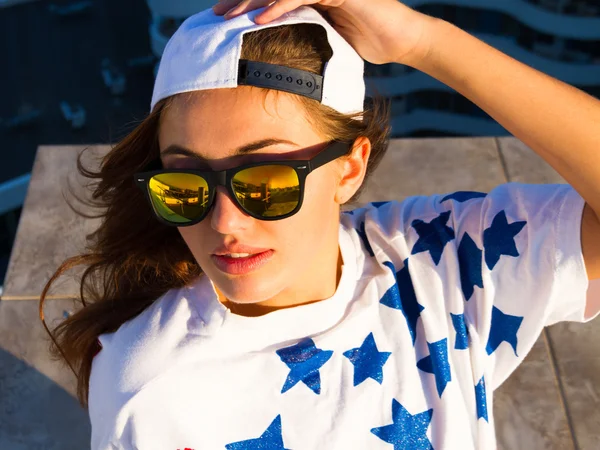 Mujer sexy joven posando en el techo, vistiendo una camiseta con estampado de bandera americana, un sombrero blanco con gafas de sol espejadas y maquillaje natural, disfrutando del atardecer y soñando. Estilo de vida retrato colores tonificados brillantes . — Foto de Stock