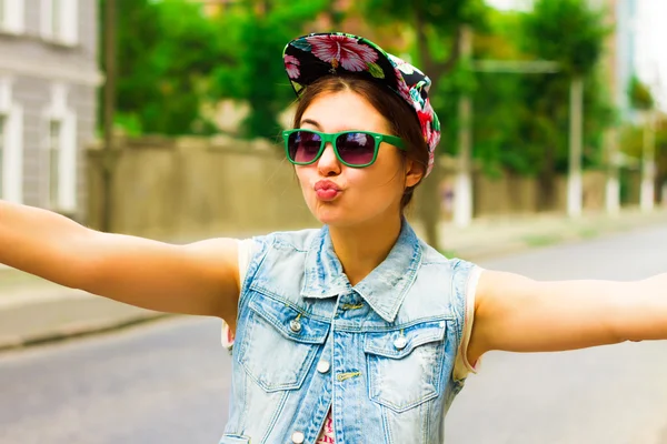 Close-up sunny portrait of pretty hipster girl having fun and want to hug. Perfect tanned skin, brunette hair. positive emotions,street background. Wearing jeans vest. floral hipster cap, sunglasses — Stock Fotó